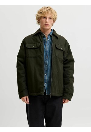 Jeune homme aux cheveux blonds bouclés portant une veste vert foncé sur une chemise en jean bleu et un pantalon noir, debout devant un fond uni.