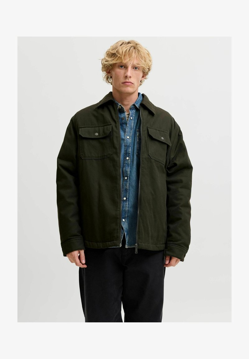 Jeune homme aux cheveux blonds bouclés portant une veste vert foncé sur une chemise en jean bleu et un pantalon noir, debout devant un fond uni.