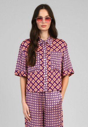 Femme aux longs cheveux bruns portant une chemise à manches courtes violette à motifs et un pantalon assorti, accessoirisée de lunettes de soleil géométriques teintées de rose.