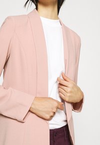 Sako v měkké růžové látce se strukturovaným designem, klopami a dlouhými rukávy, vrstvené přes bílou košili, s minimálním zdobením.