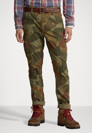 Vihreät camo cargo-housut, joissa on moniväristä hyönteiskuosittua brodeerausta, sivutaskut ja käänteet, yhdistettynä ruskeisiin nauhakenkiin.