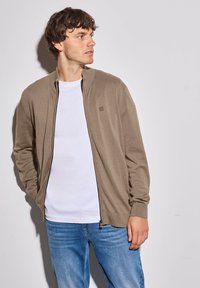 Béžový svetr na zip s vysokým límcem, žebrovanými manžetami a spodním lemem. Nositý přes bílý tričko a v kombinaci s modrými džíny.