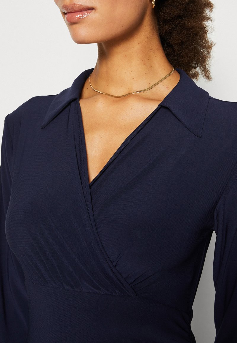 Marineblaues Wickelkleid mit einem breiten Kragen, das aus weichem, glattem Stoff und subtilen Faltendetails besteht. Eine goldene, geschwungene Halskette betont den Ausschnitt.