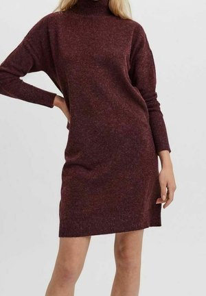 Donna che indossa un vestito maglione bordeaux scuro, a maniche lunghe e lunghezza al ginocchio, con collo alto, in piedi con una mano sul fianco.