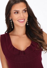 Burgundi ujjatlan felső fodros dekoltázzsal, bordás textúrával. Hosszú hajú modell, lógó virág fülbevalóval. Világos háttér.