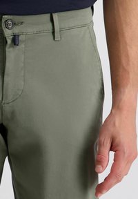 Helle olivgrüne Hosen mit glatter Textur, mit einem vorderen Knopfverschluss und seitlichen Taschen, vor einem neutralen Hintergrund präsentiert.