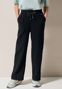 Pantalons noirs texturés avec une coupe ample, dotés d'une taille élastique et d'un cordon de serrage. Associés à des chaussures de sport légères.