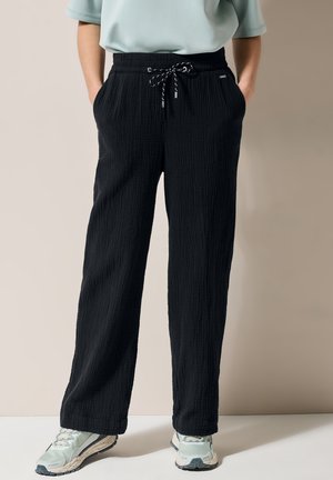 Pantalons noirs texturés avec une coupe ample, dotés d'une taille élastique et d'un cordon de serrage. Associés à des chaussures de sport légères.