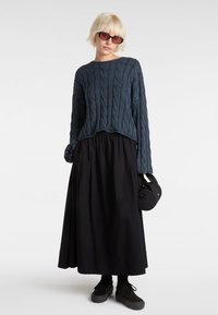 Oversized cable-knit sweater in dark blue, paired with a long black skirt. Black sneakers and a small round bag complete the look.