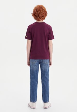 Barn med krøllet rødt hår iført en mørkerød t-shirt, blå jeans og hvide sneakers står med ryggen til på en ensfarvet hvid baggrund.