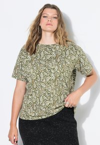 Zelená vzorovaná blůza s krátkým rukávem, která má paisley design s bílými a oranžovými akcenty. Měkká látka s volným střihem, nošena s černou sukní.