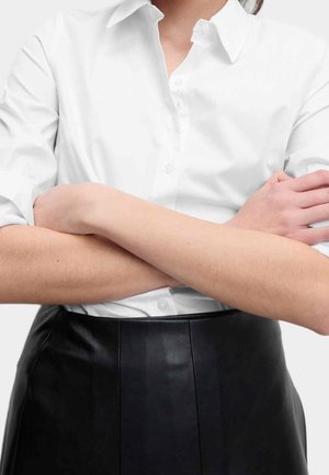 Personne portant une chemise blanche boutonnée et une jupe noire, les bras croisés sur la poitrine, visible du cou à la taille.