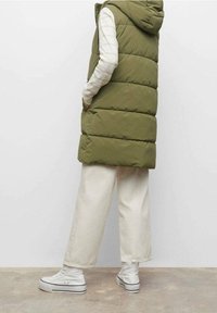 Oliwkowa pikowana kamizelka z kapturem, z przeszywanym wzorem i bocznymi kieszeniami, noszona na białej, paskowanej bluzce z długim rękawem i kremowych spodniach.