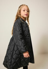 Geacă puffer neagră matlasată, cu guler rotund, mâneci lungi și o formă A-line avantajoasă. Include un fermoar frontal și un model în diamant.