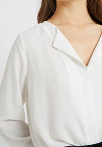 Blusa bianca realizzata in un tessuto morbido e leggero. Presenta un colletto a V, maniche lunghe e una lucentezza sottile. Texture liscia senza motivi visibili.