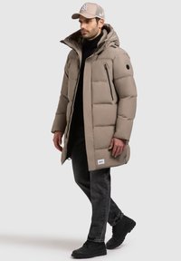 Palton lung căptușit bej cu glugă, prezentând fermoare negre și un patch cu logo. Asortat cu blugi închisi la culoare și ghete negre, purtat de un model masculină.