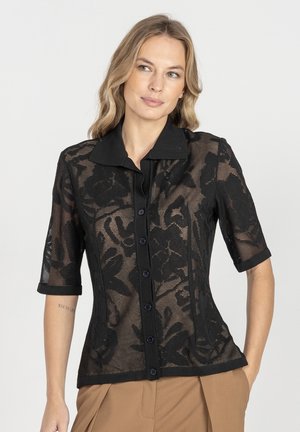 Kobieta z falującymi blond włosami, ubrana w czarną przezroczystą koronkową bluzkę z kwiatowym wzorem i beżowe spodnie, stojąca z rękami częściowo w kieszeniach na białym tle.