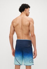 Costume de baie cu un gradient albastru închis și verde teal, cu un finisaj texturat, având un buzunar la spate și detalii de contur contrastante.