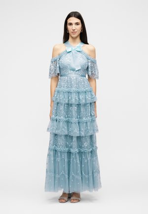 Femme debout portant une robe longue en dentelle bleu clair avec épaules dénudées, volants superposés et un nœud au niveau de l'encolure, accompagnée de sandales.