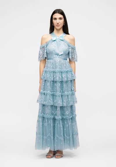 Femme debout portant une robe longue en dentelle bleu clair avec épaules dénudées, volants superposés et un nœud au niveau de l'encolure, accompagnée de sandales.
