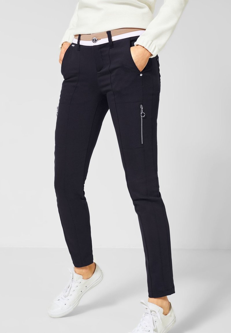 Pantalon noir slim avec fermetures éclair latérales, poches et finition texturée. Associé à des baskets blanches basses pour un look décontracté.