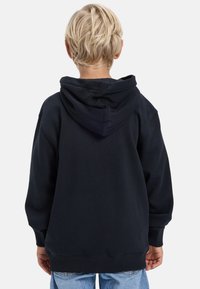 Dieťa s krátkymi blond vlasmi, oblečené v čiernej mikine a modrých džínoch, otočené chrbtom na jednoduchom bielom pozadí.