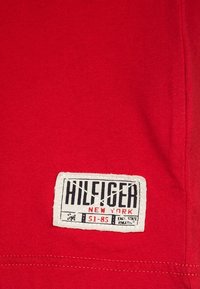 Punainen puuvillakangas, jossa on selkeästi näkyvissä etiketti, jossa lukee "HILFIGER" ja "NEW YORK" lihavalla mustalla tekstillä valkoisella taustalla.