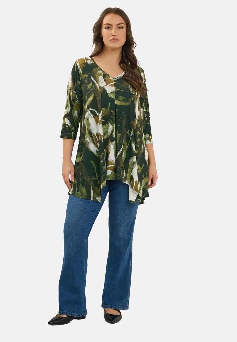 Bloemen groene tuniek met driekwart mouwen, V-hals en asymmetrische zoom, gecombineerd met blauwe uitlopende jeans en zwarte ballerina's.