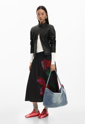 Nő fekete bőrdzsekit és fekete, piros mintás szoknyát visel, nagy kék farmer táskát tart a kezében, piros tornacipő van rajta, fehér háttér előtt áll.