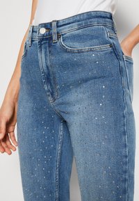 Jeans de mezclilla de color azul claro con cintura alta, adornados con tachuelas metálicas dispersas, bolsillos delanteros y un diseño clásico de cinco bolsillos.