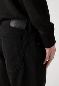 Des jeans en denim noir présentent une étiquette en cuir au niveau de la taille. Gros plan sur la poche arrière avec des détails de couture subtils.