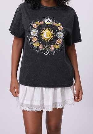 Person trägt ein schwarzes T-Shirt mit Sonnen-, Mond- und floralem Himmelsdesign, kombiniert mit einem weißen Rock mit Ösen-Spitzenbesatz.