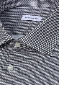 Chemise habillée à col avec un petit motif géométrique noir et blanc et des boutons blancs, étiquetée "seidensticker" à l’intérieur du col.