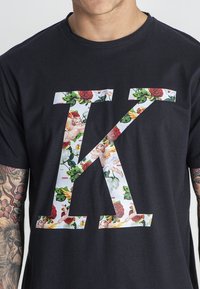 Černé tričko s velkým, velkým písmenem "K" vyplněným barevným květinovým vzorem a cheruby. Krátké rukávy, kulatý výstřih, bavlněný materiál.