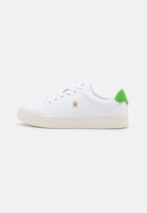Zapatilla blanca de corte bajo con suela beige, lengüeta verde en el talón, cierre de cordones en la parte delantera y pequeño logo dorado "TH" en el lateral.