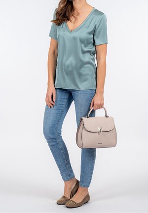 Mujer con blusa de satén verde azulado con escote en V, vaqueros ajustados azul claro y zapatos planos marrones con tachuelas, sosteniendo un bolso beige a su lado.