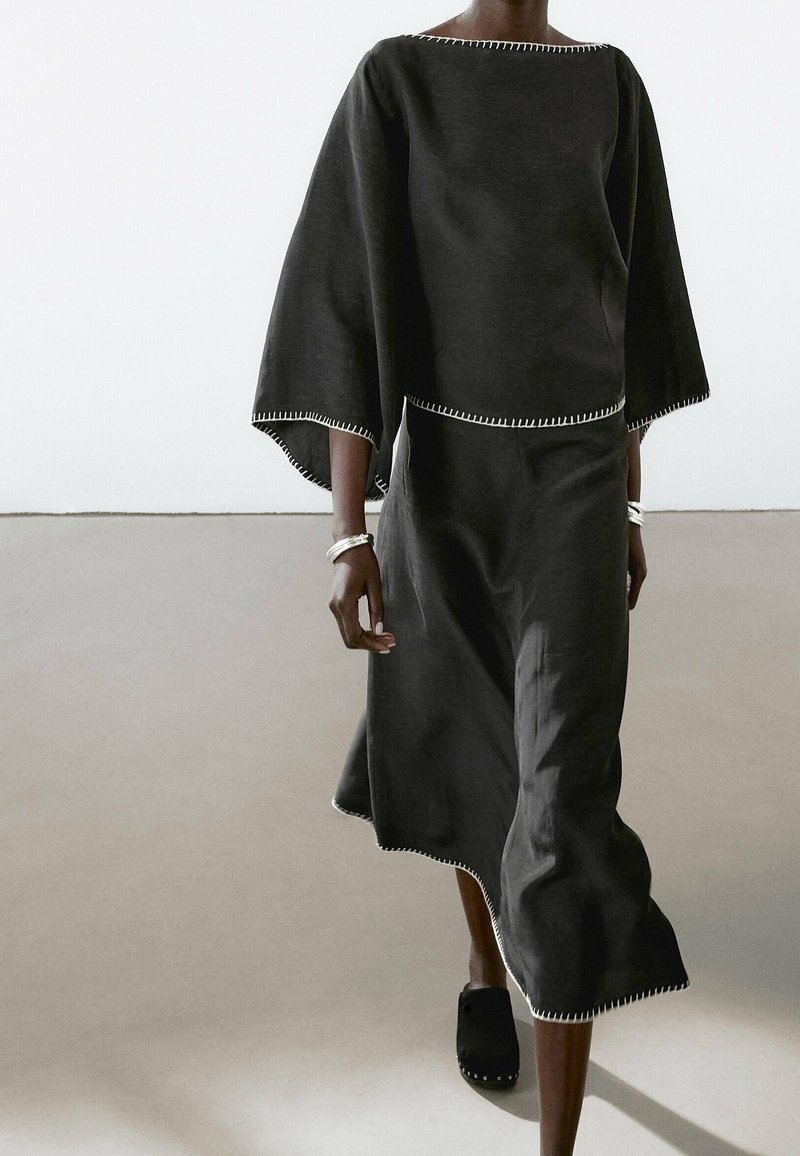 Femme portant une robe noire ample avec de larges manches et des détails de couture blanche, associée à des sabots noirs assortis et des bracelets argentés.