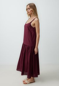 Abito maxi senza maniche in malva con corsetto aderente e gonna arricciata. Realizzato in tessuto leggero, presenta spalline sottili e una silhouette fluida.