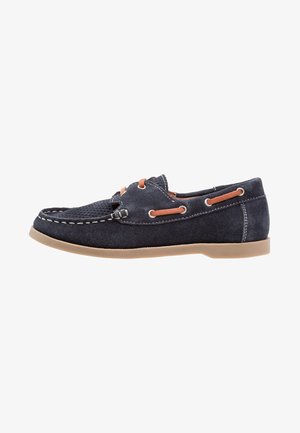 Scarpa da barca in camoscio blu navy con punta perforata, cuciture a contrasto, occhielli beige e suola in gomma. Presenta un design in stile mocassino.