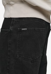 Svarta jeans i denim med en bakficka som har en liten etikett. Tyget har en lätt struktur och kompletteras av en ljusfärgad sweatshirt.