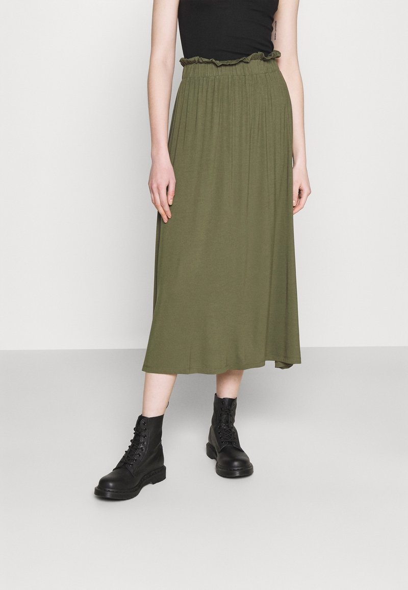 Femme portant une jupe midi vert olive avec une taille élastique, associée à des bottes de combat noires lacées, sur un fond uni.