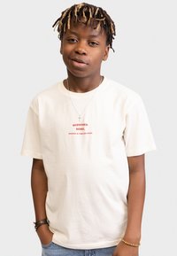 Light beige cotton t-shirt with short sleeves, featuring red text that reads "REDEFINED REBEL" and "PROSPER IN TIMELESS SPACE."
