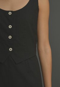 Top en lin noir avec un col rond, cinq boutons sur le devant avec une finition en bois clair, et un ourlet asymétrique.