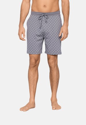 Miehen vartalo ja jalat, joissa on harmaat, kuviolliset nyörishortsit, seisoo paljain jaloin yksinkertaista vaaleaa taustaa vasten.