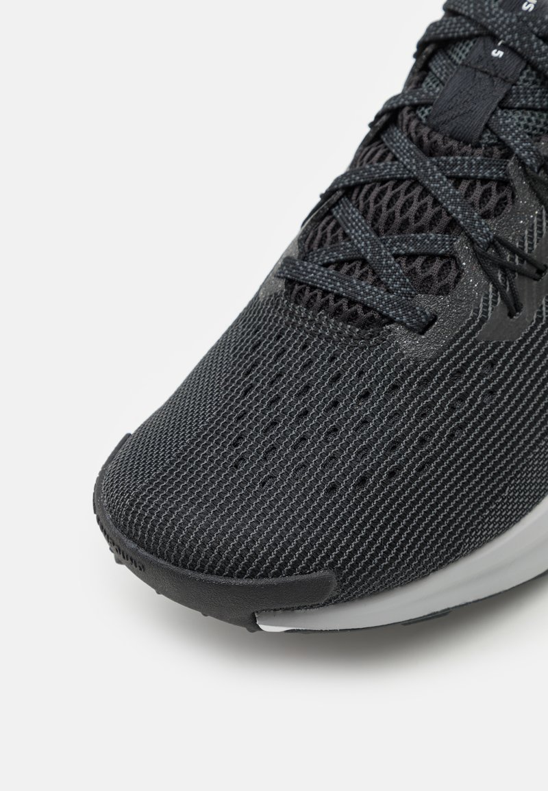 Zapatilla deportiva negra con parte superior de malla texturizada, con acentos grises, costuras intrincadas y una suela de goma suave. Sistema de lazado detallado.