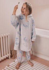 Peignoir à capuche en peluche bleu clair avec des imprimés de dessins animés ; pantalon de pyjama gris avec des motifs assortis. Le sol est recouvert d'un tapis texturé.