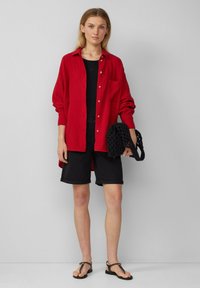 Rotes Hemd mit Knopfleiste und Tasche, schwarzes Tanktop, schwarze hochgekrempelte Shorts und schwarze geflochtene Tasche. Sandalen runden das Outfit ab.