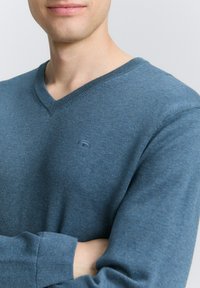 V-Ausschnitt Pullover aus blauem Strickstoff, mit gerippten Bündchen und Saum, ausgestattet mit einem kleinen Logo auf der linken Brustseite. Glatte Textur.