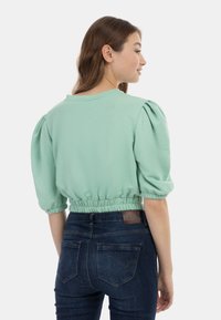 Menta zöld cropped top puffos ujjal és ráncolt aljjal, sötétkék farmernadrággal párosítva, amelyen bőrdíszítés található a hátsó részen.