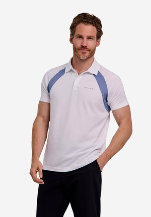 Mann mit kurzem lockigem Haar, der ein weißes Polohemd mit blauen Schulterpaneelen und schwarze Hose trägt, steht mit einer Hand in der Tasche.